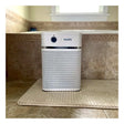 An Austin Air Systems HealthMate Junior Machine A200, featuring a sleek white design, sits on a beige textured rug in a tiled bathroom. Positioned in front of a bathtub with decorative trim and a window in the background, this medical-grade HEPA air purifier seamlessly complements the tranquil space while providing outstanding air quality.