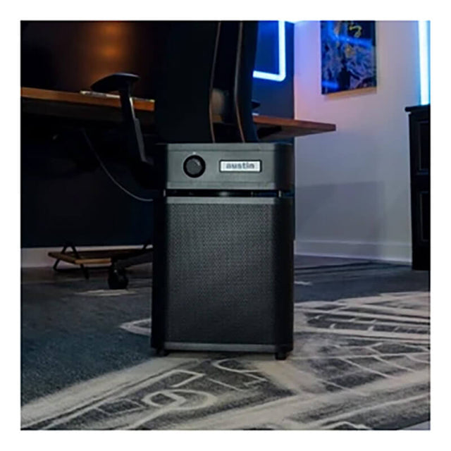 An Austin Air Systems HealthMate Plus Junior Machine A250 in black, featuring medical-grade HEPA filtration and advanced VOC removal, sits on a carpeted floor in a modern office with a desk, chair, neon lights, and wall art in the background.