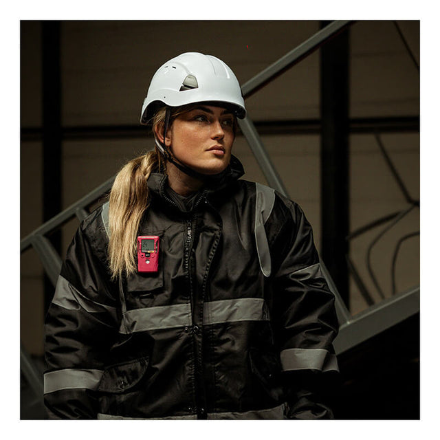 A person wearing a white hard hat, black reflective safety jacket, and a pink WatchGas SST1 2 Year H2S Gas Detector (model SST1-H-24-P) stands indoors near metal stairs, looking to the side.
