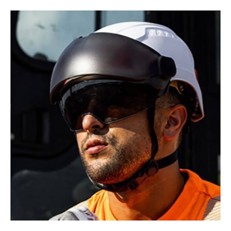 A man wearing a PIP Traverse Eyewear Protector 251-HP1491P and a white safety helmet with black visor, dressed in an orange hi-vis shirt, stands outdoors in bright sunlight.